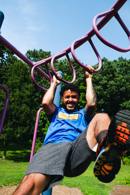 A person wearing a blue t-shirt with a graphic design in the center, hanging on monkey bars. 