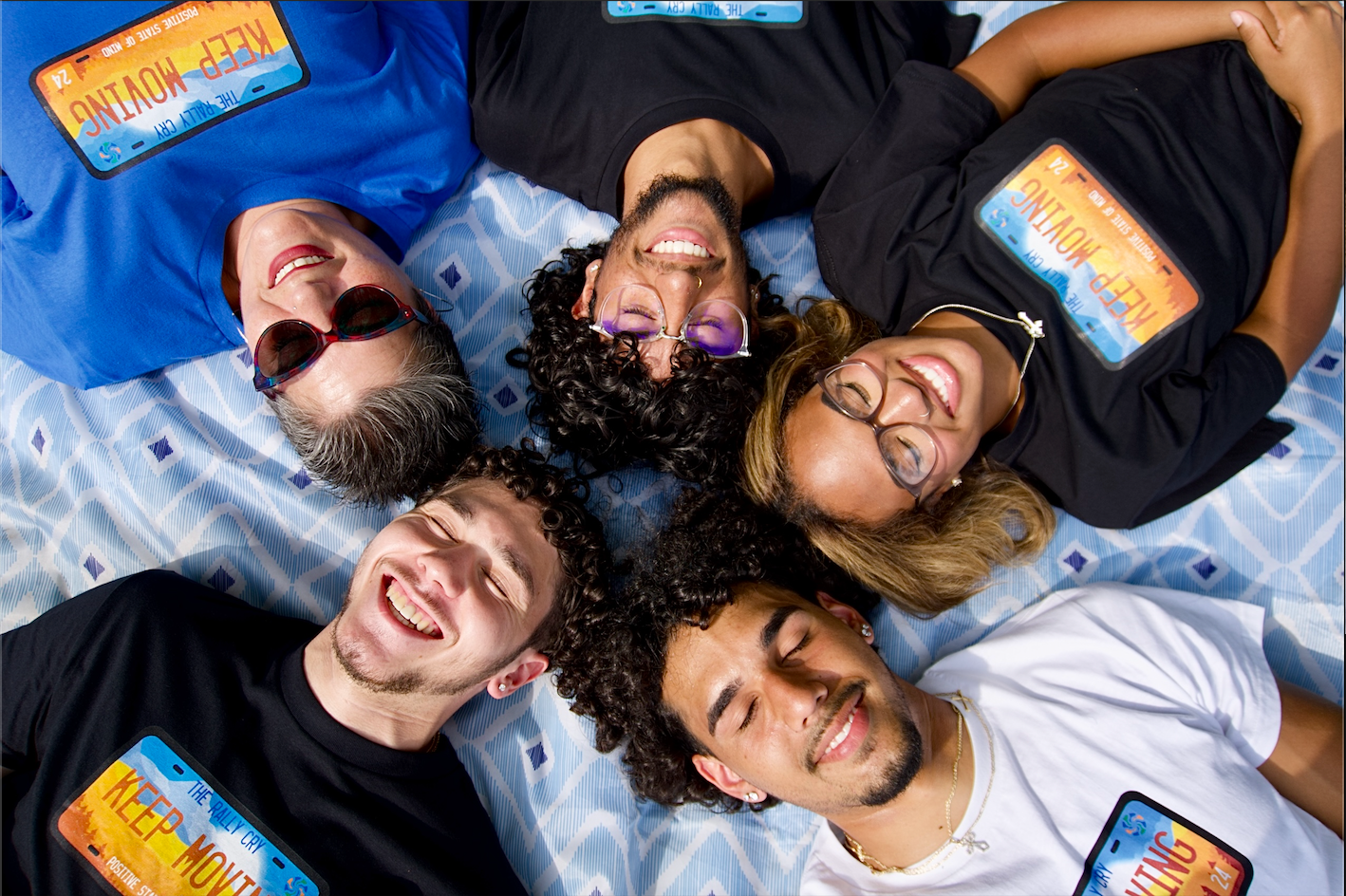 three men and two woman laying down in a circle smiling with wearing blue, white, and black keep moving tees